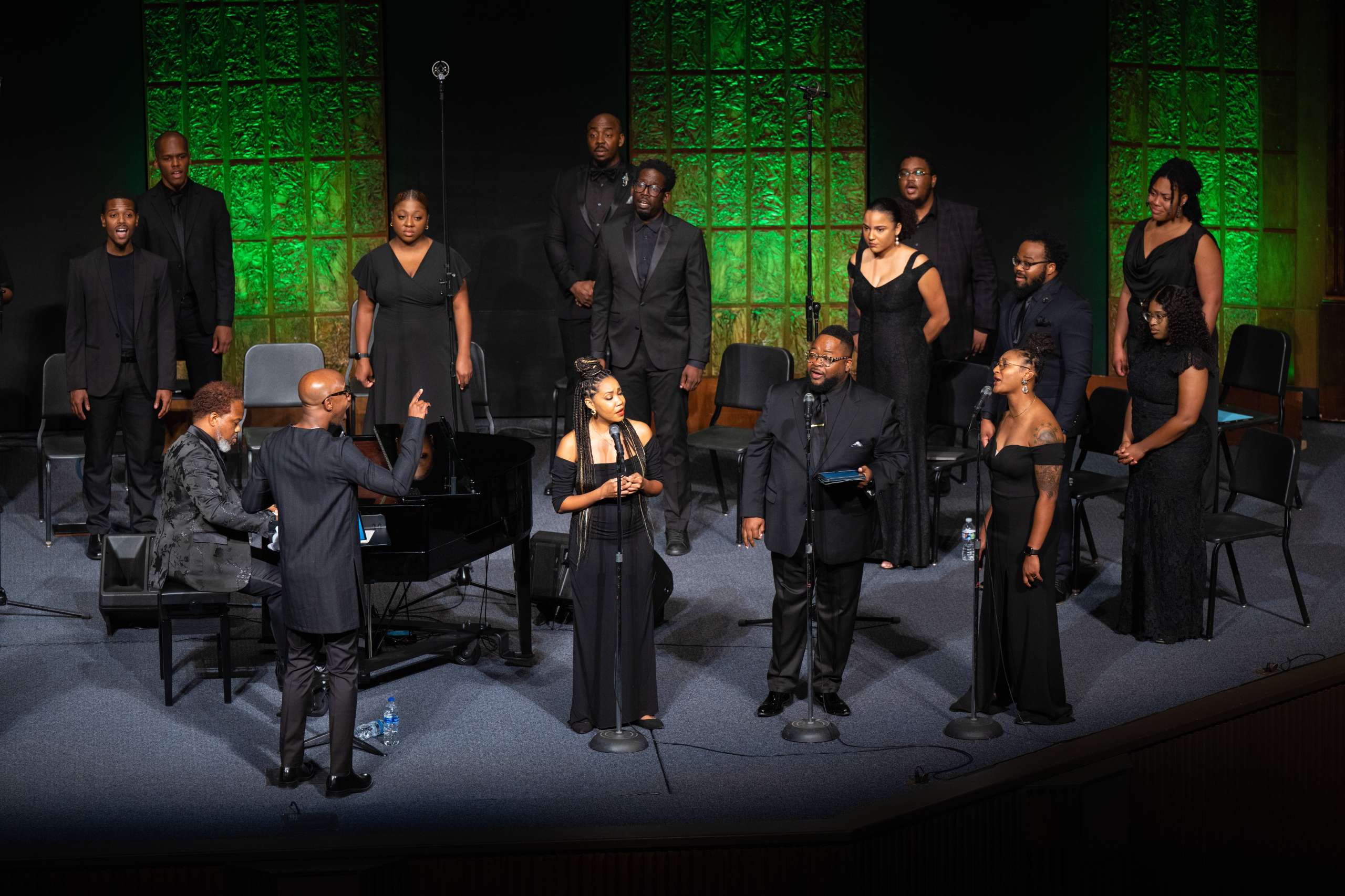 An ensemble of singers dressed in formal black attire stands ready to perform on stage, with a male conductor at the forefront, gesturing expressively towards a seated pianist.