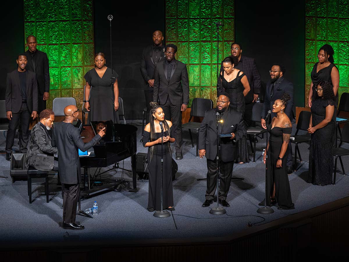 A choir of elegantly dressed individuals stands on stage ready to perform, with some members positioned by microphones. A conductor stands in the foreground, directing a pianist or another musician seated at a grand piano.